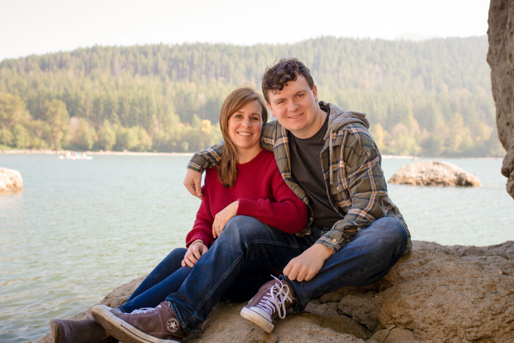 Mom and teenage son are sitting on a rock at Rattlesnake Lake. Teenage son has his arm around his mother.
