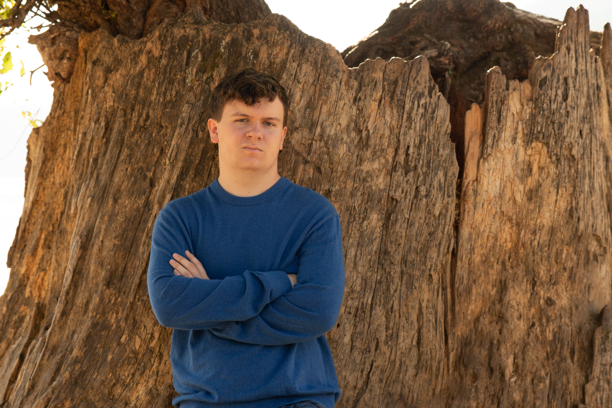 A young man with short dark hair stands in front of a large, rugged tree trunk or wood structure, wearing a blue long-sleeve sweater with arms crossed. He gazes at the camera with a serious expression, and the natural outdoor setting provides a warm, earthy backdrop.