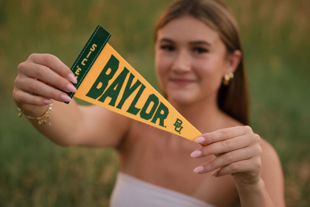 High school senior smiling while holding a Baylor University pennant during an outdoor portrait session, photographed in a natural Colorado setting for Denver Senior Photography.