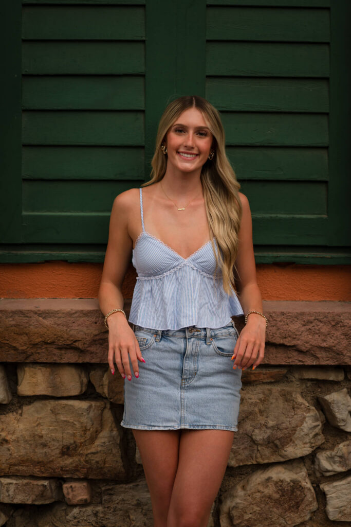 Senior girl in a striped top and denim skirt posing in front of green shutters during a Castle Rock senior photo session
