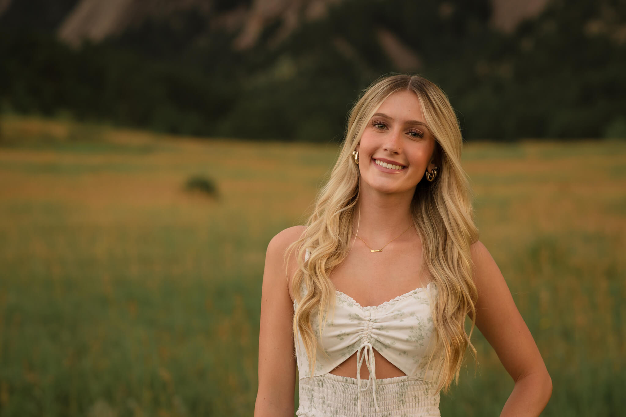 Smiling high school senior in a floral top standing in a grassy field with mountains in the background during a Castle Rock senior photo session