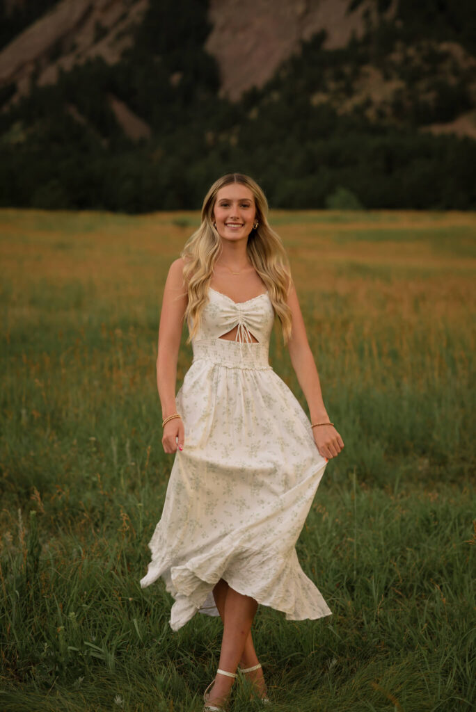 High school senior in a flowy white dress walking through a grassy field with mountain views during a Castle Rock senior photo session