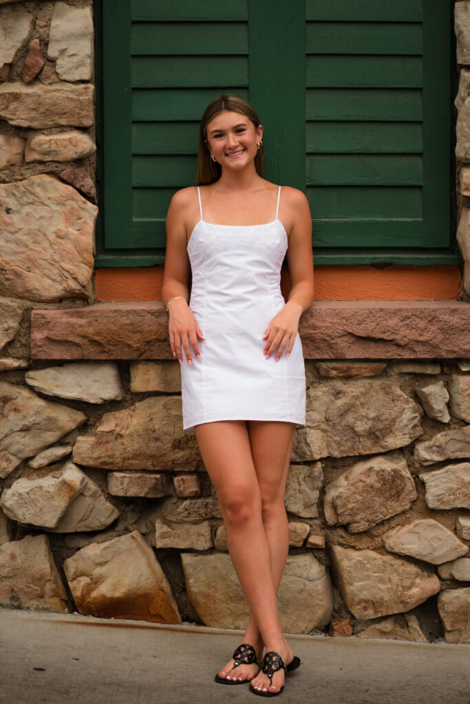 Senior girl in a white dress posing against a stone wall and green shutters during a Castle Rock senior photo session