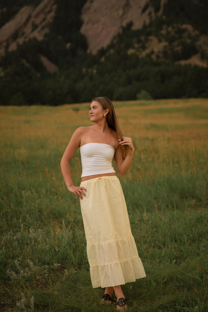 High school senior in a yellow skirt and white top posing in a grassy field with mountains during a Castle Rock senior photo session
