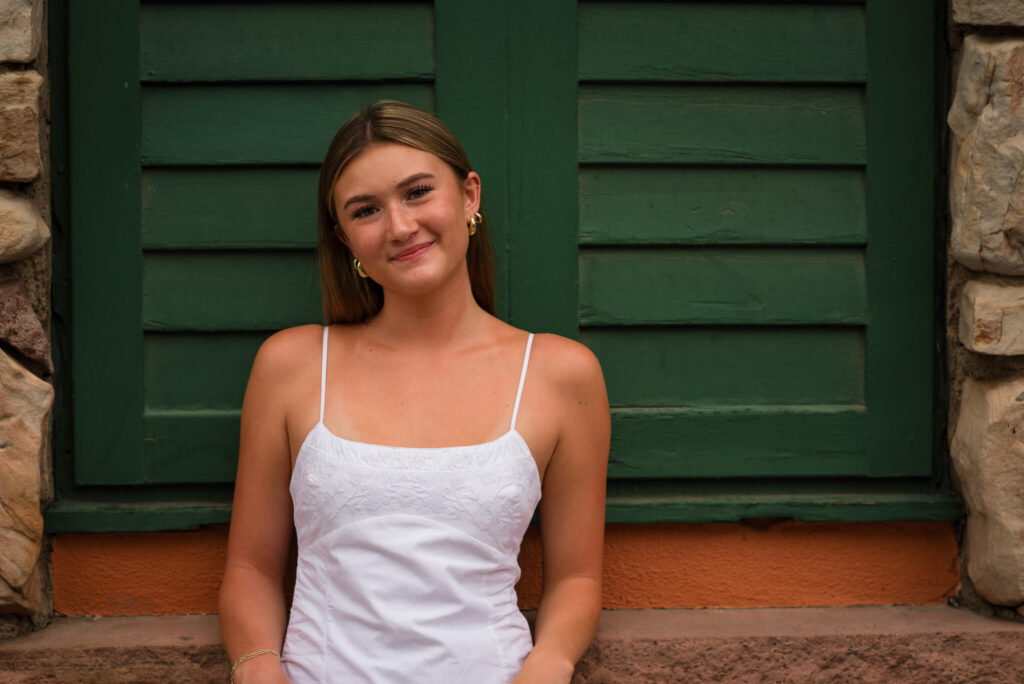 High school senior girl in a white dress posing in front of a green wooden wall during an urban Denver senior photo session.