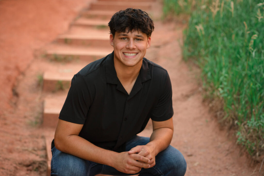 Smiling high school senior guy sitting on red rock steps during an outdoor Denver senior photo session with natural Colorado scenery.
