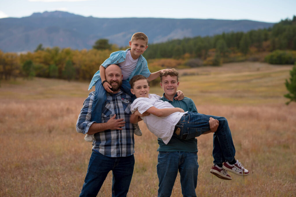 Father carrying his sons in a grassy Colorado field with mountain views during a fun outdoor family photo session.