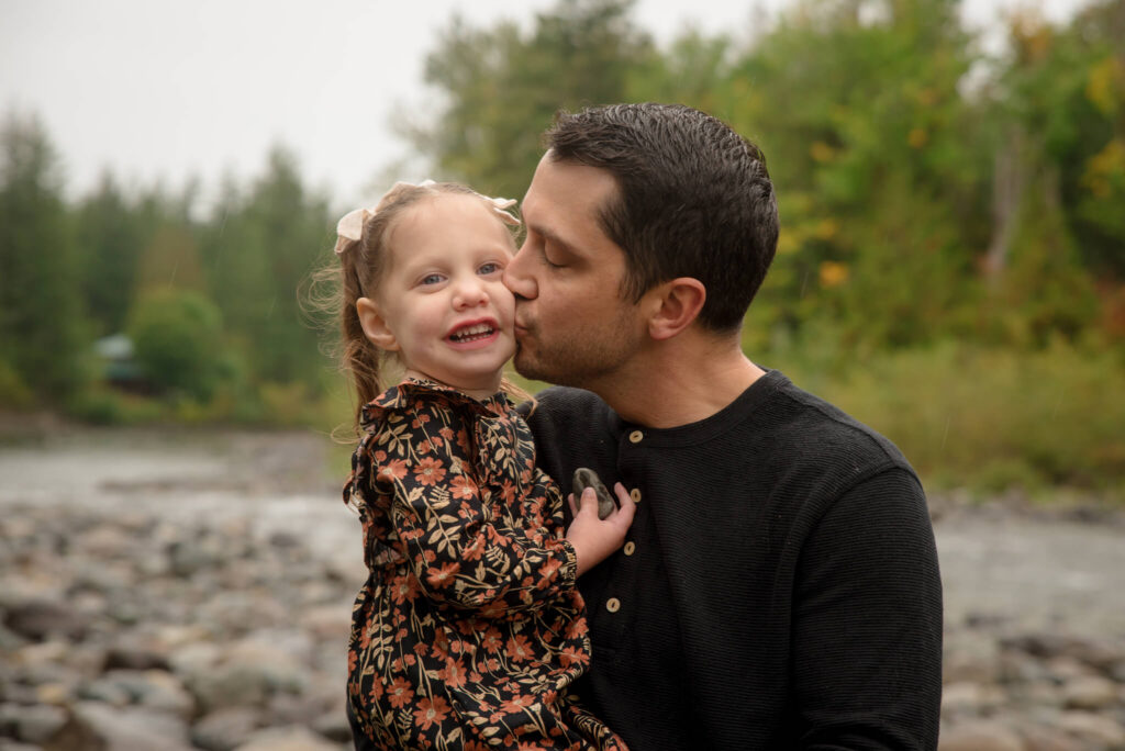 Father kissing his young daughter on the cheek beside a river during a heartfelt outdoor family photography session in Colorado.