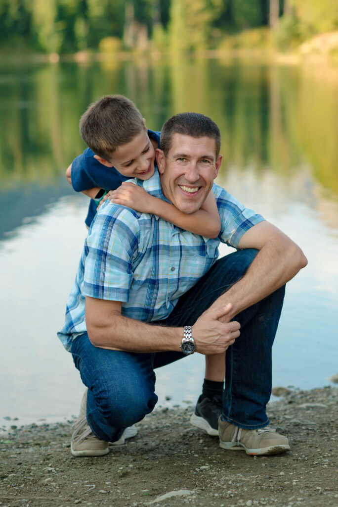 Smiling father kneeling by a lake while his young son hugs him from behind during a candid Colorado family photography session.