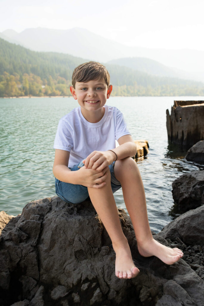 Boy sitting on lakeside rocks with mountain views behind him during an outdoor summer family photography session.