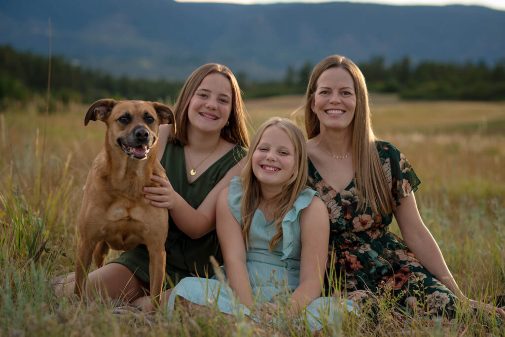 Mother and daughters sitting in a grassy field with their dog during a summer family photo session in Colorado.