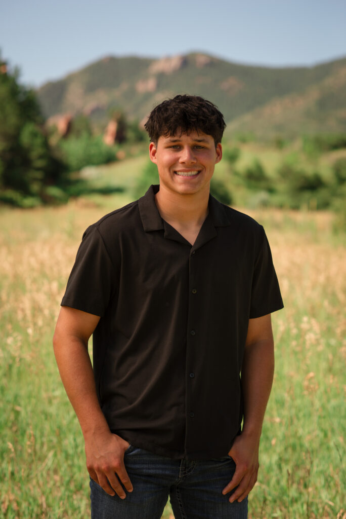 Senior boy posing in a grassy Colorado field with beautiful mountain and red rock views in the background.