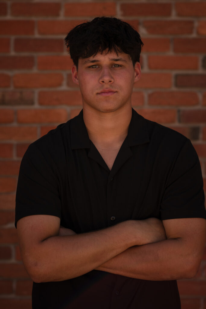 Colorado senior pictures for guys featuring a senior boy in a black shirt posing with crossed arms against a brick wall.