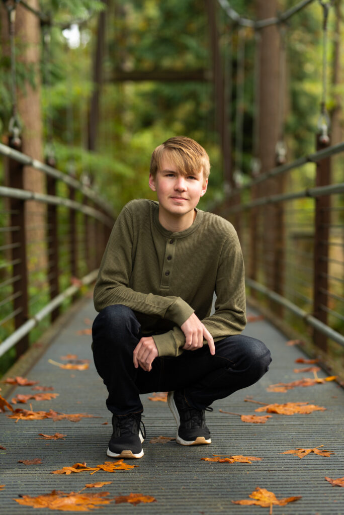 Colorado senior pictures for guys featuring a senior boy crouching on a wooded bridge surrounded by fall leaves.