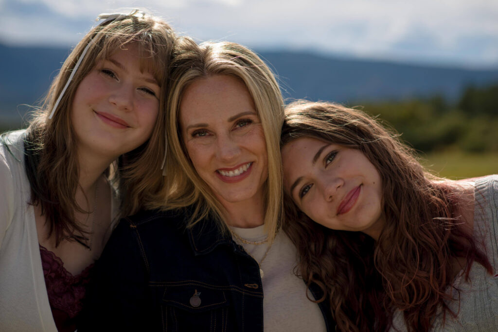Mom and her two daughters cuddled together and smiling during a fall family mini session in Castle Rock, Colorado, with open space and mountain views in the background.