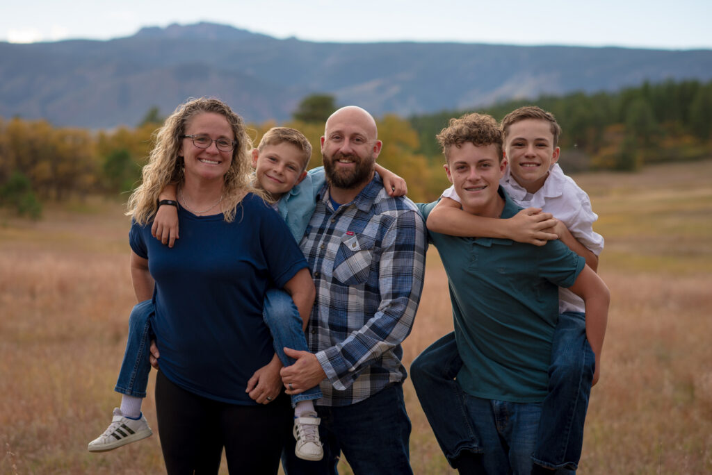 Family of five smiling and having fun together in a golden field with Colorado mountain views during a fall family mini session in Castle Rock, Colorado.