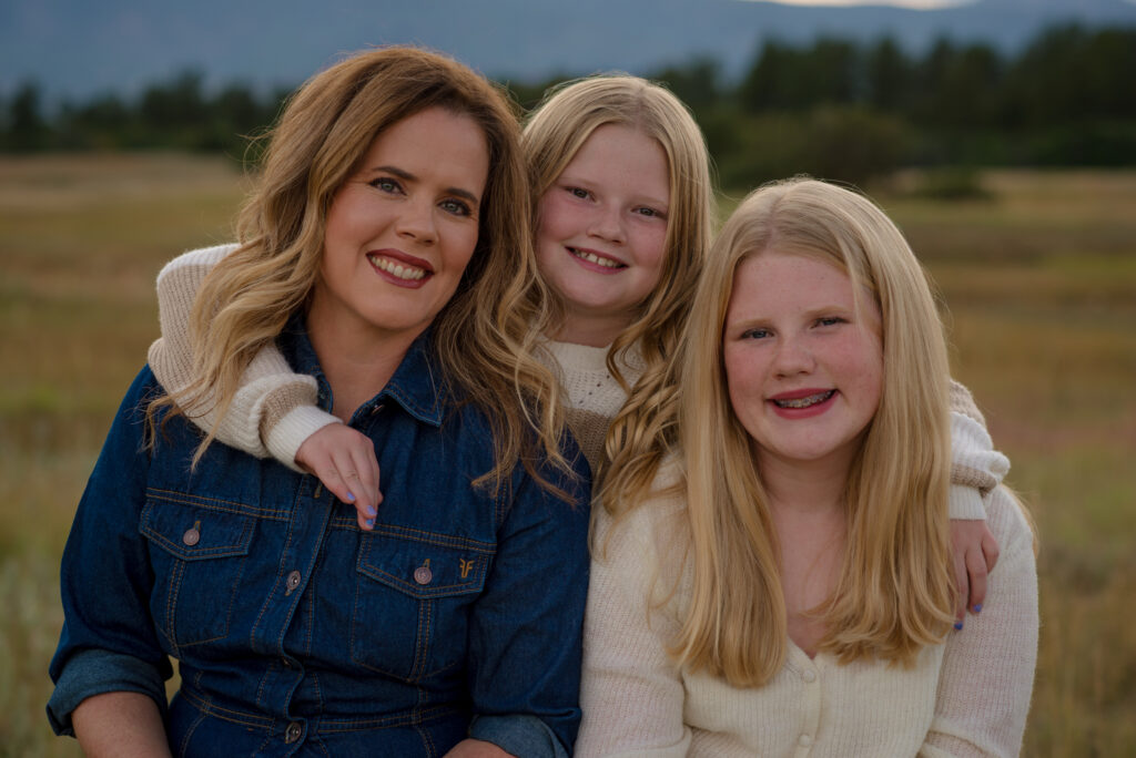 Alt text: Mom and her two daughters smiling together during a fall family mini session in Castle Rock, Colorado, with beautiful open space and mountain views in the background.