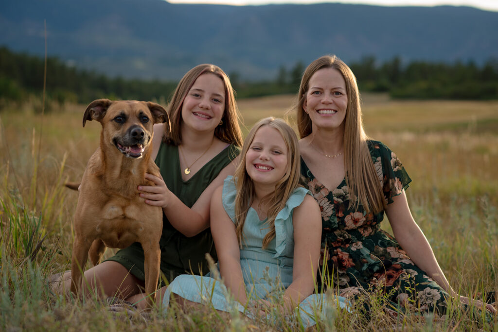 Mom and her two daughters smiling with their family dog during a fall family mini session in Castle Rock, Colorado, surrounded by golden grasses and mountain views.