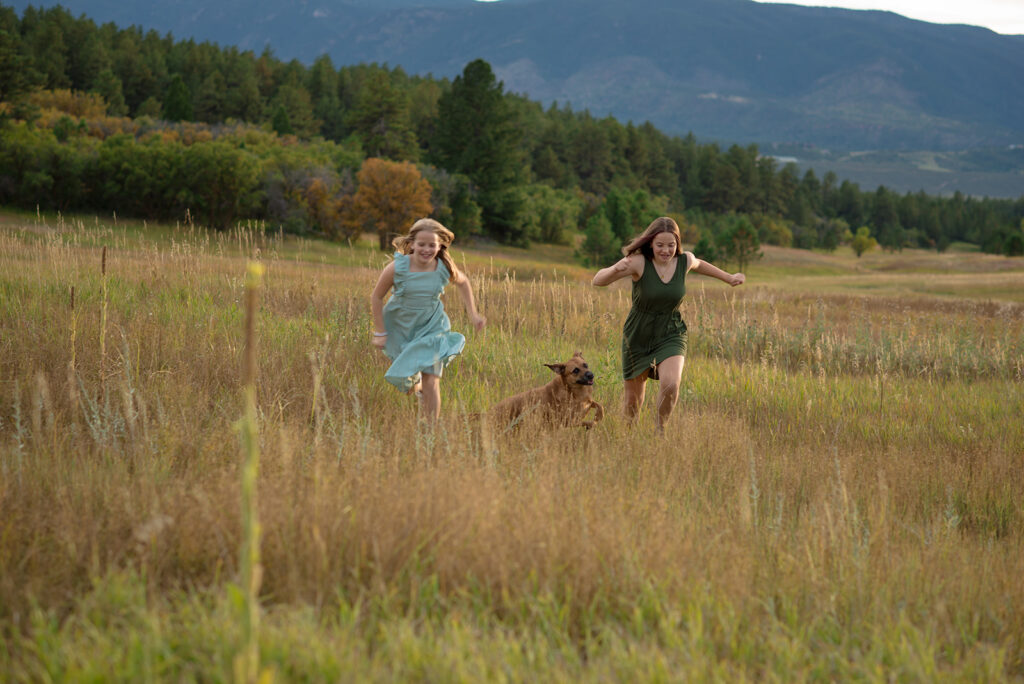 Two sisters running through golden fall grasses with their family dog during a fall family mini session in Castle Rock, Colorado, with mountain views in the background.