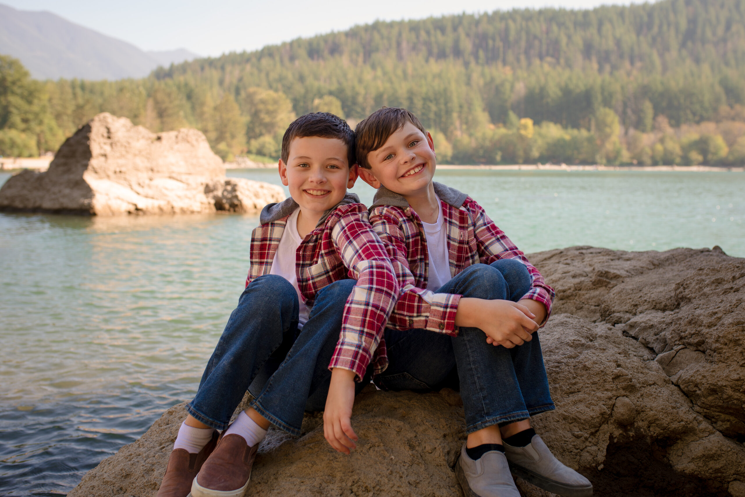 Portrait captured by a Castle Rock photographer of two young boys sitting on a rock by a lake, smiling and hugging each other while wearing matching plaid shirts, with mountains and a forested shoreline in the background.