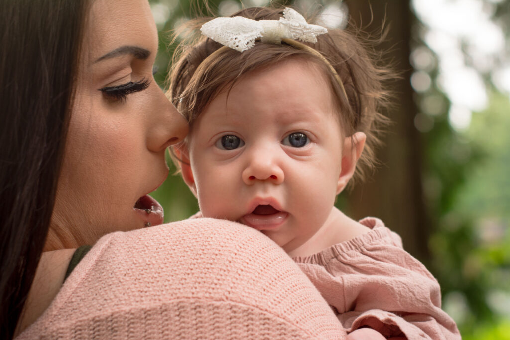 A mother is holding her baby girl on her shoulder and whispering in her ear.  Baby girl is looking at the camera.  Mom and baby girl are at Pine Lake Park in Sammamish, WA.