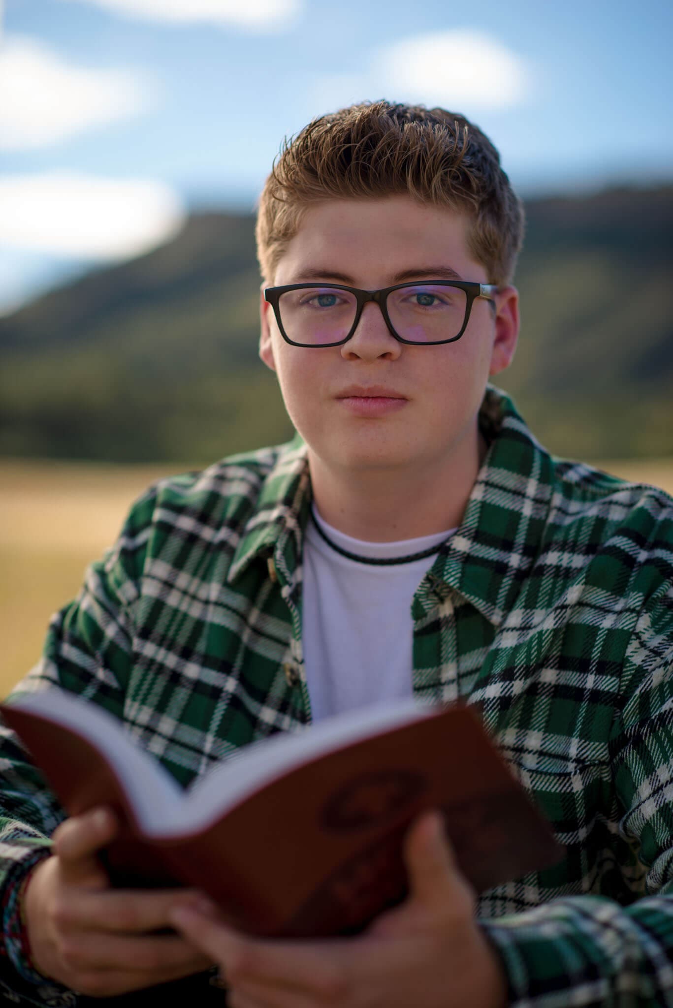 High school senior portrait of a student wearing glasses and a green flannel, holding a book in a scenic outdoor Colorado setting, captured by a Colorado Senior Photographer.
